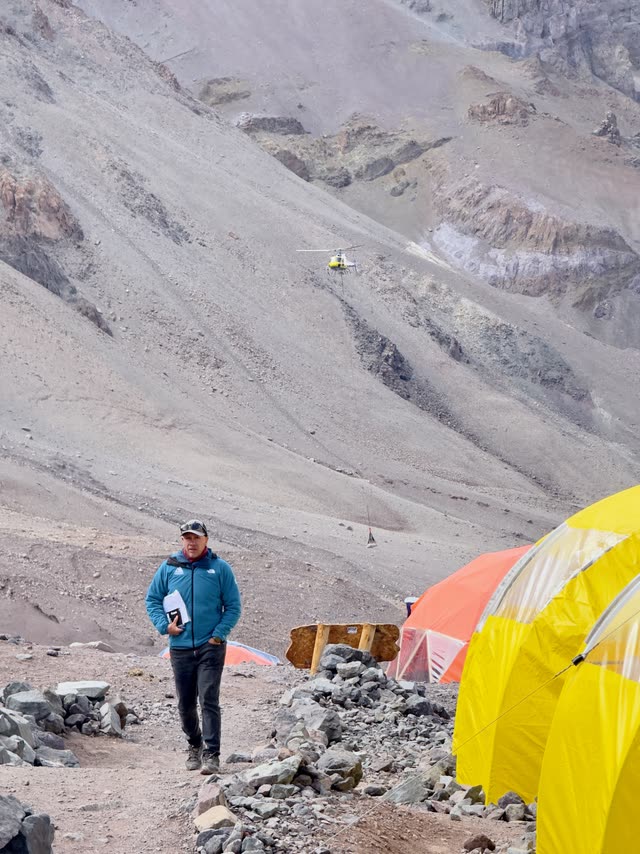 Person in mountaineering gear near tents with helicopter in background