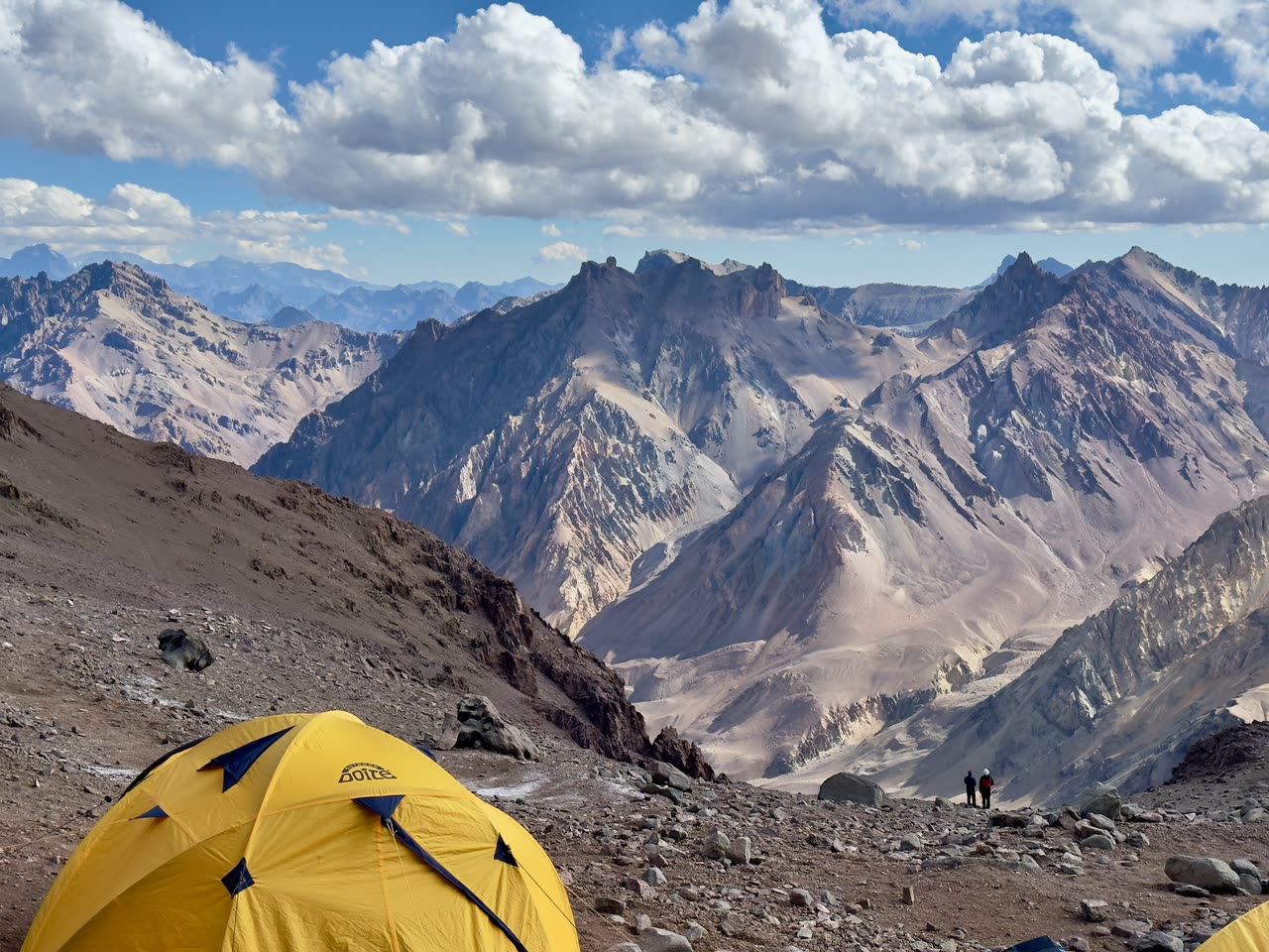 Yellow tent on steep rocky slope with snowy peaks beyond
